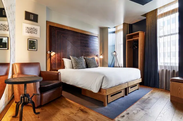 A king bed with white linen and a leather headboard sits beside a leather armchair, with an open wardrobe and tall windows in a hotel bedroom.