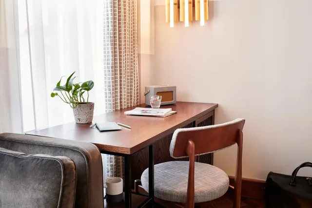 A wooden desk with a chair, a potted plant, a glass and a notebook stands by sheer curtains, with a grey armchair in the foreground.