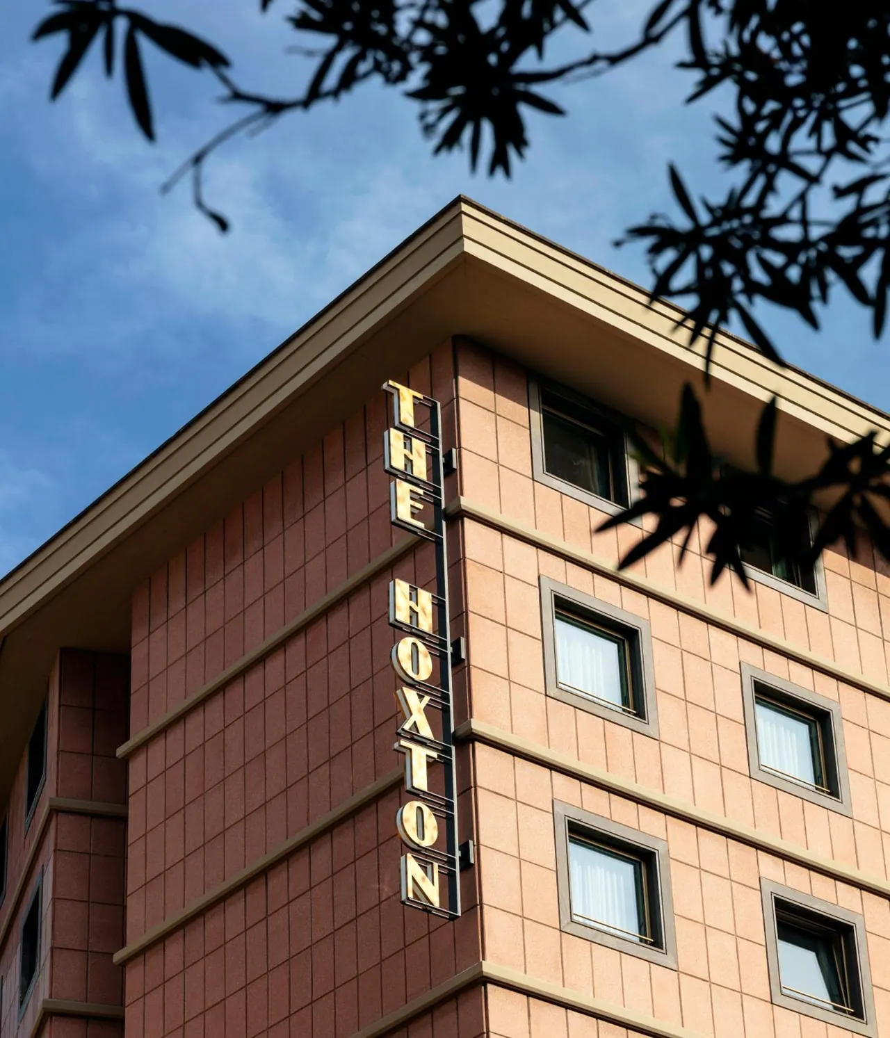 Looking up at the tiled exterior of a building with a vertical sign reading THE HOXTON against a blue sky, with tree branches in the foreground.