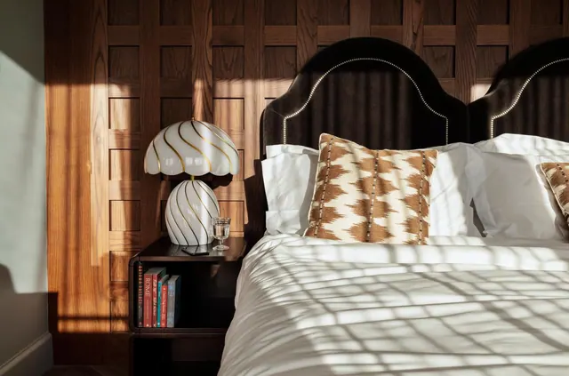 A made bed with white linen and a patterned cushion sits beside a bedside table holding a striped table lamp, books and a glass of water, against a wood-panelled wall.