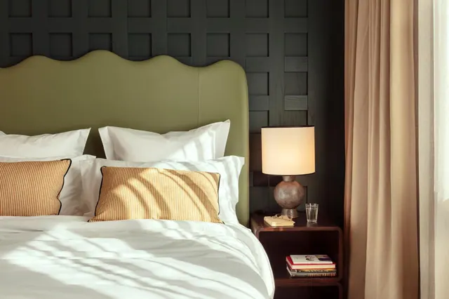 A made bed with white linens, an olive green upholstered headboard, striped cushions, and a lamp on a dark wooden bedside table.