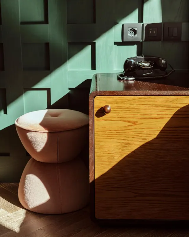 Two stacked pink upholstered poufs stand beside a wooden cabinet topped with a black rotary telephone, in front of a green paneled wall lit by sunlight.