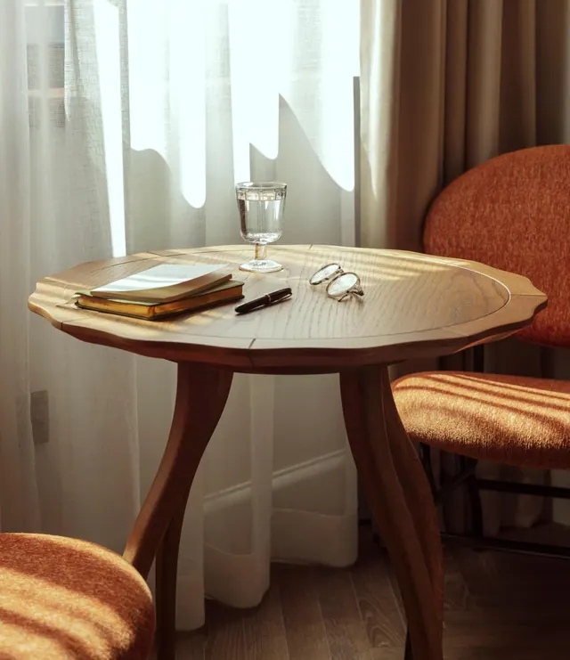 A round wooden side table by a sheer-curtained window holds a glass of water, notebooks, a pen and reading glasses, with orange upholstered chairs beside it.