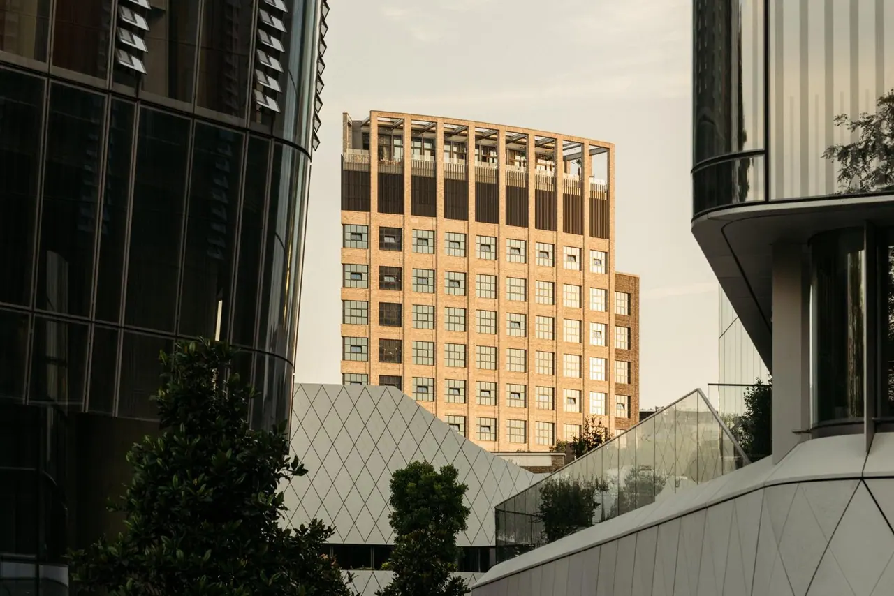Exterior view between modern glass towers, showing a brick building with open upper terraces and angular panelled facades in the foreground.
