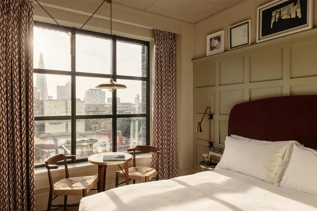 A bedroom with a made double bed, a small round table with two chairs by a window, and a view over city rooftops to The Shard.