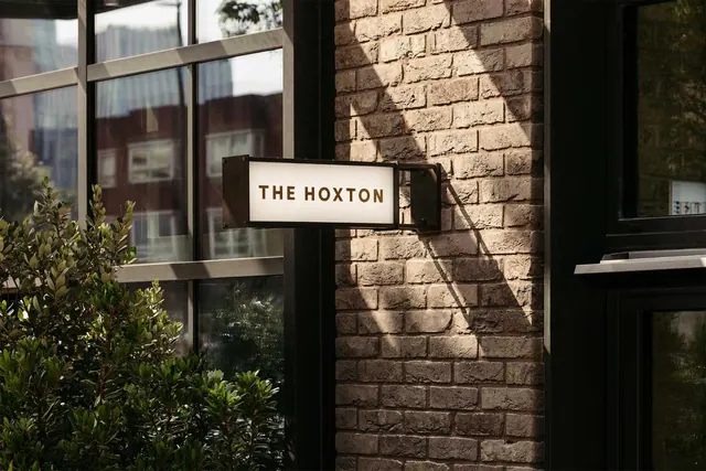 A illuminated projecting sign reading THE HOXTON hangs on a brick wall beside large glass windows and green shrubs.