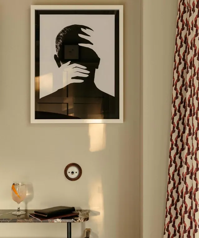 A framed black and white silhouette print hangs above a marble console table holding a drink and notebooks, beside a patterned curtain.