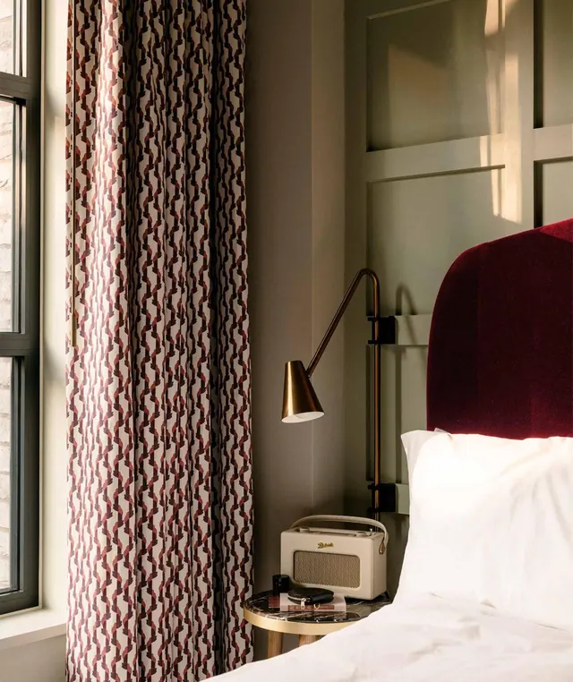A bedroom corner shows a made bed with a dark red upholstered headboard, a brass wall lamp, a cream radio on a small side table and patterned curtains beside a window.