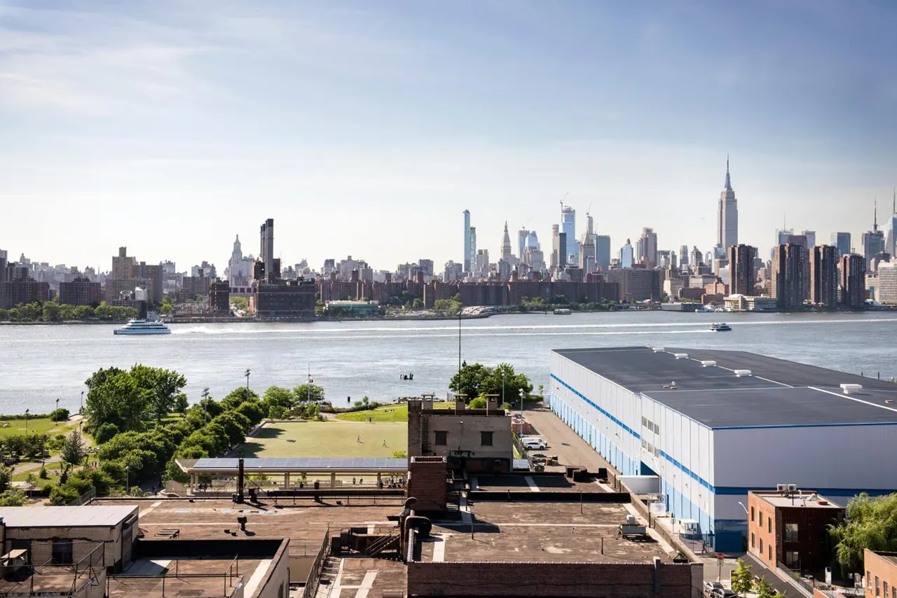 Elevated view over a river toward the Manhattan skyline, including the Empire State Building, with rooftops, a park and a large industrial building in the foreground.
