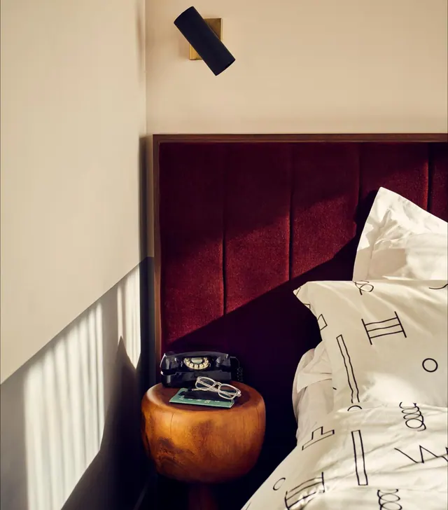 A bedroom with an upholstered burgundy headboard, a black rotary telephone and glasses on a wooden side table, and white bed linen with black line patterns.