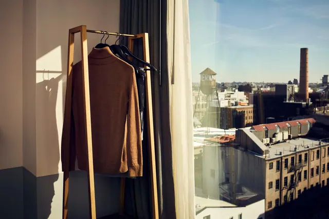 A brown knit turtleneck and dark garments hang on a wooden rack beside a window that looks out over brick buildings, a smokestack and a water tower.