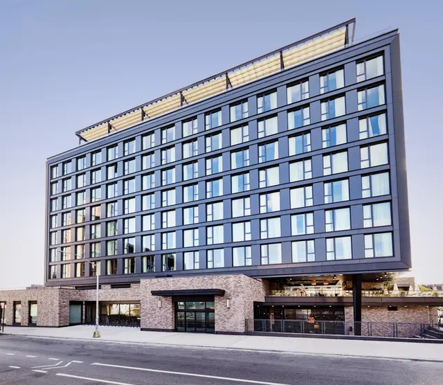 Exterior view of a multi-storey hotel building with a dark grey facade, rows of windows, a brick ground floor and an empty street in the foreground.