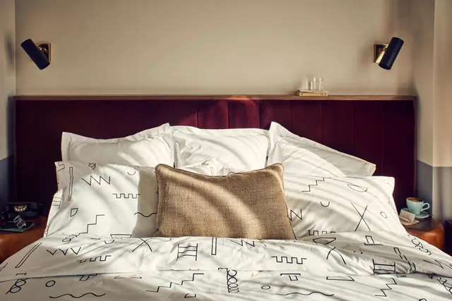 A made bed with white linen printed with black line doodles, a tan cushion and a brown velvet headboard, with wall lights and a cup on the bedside table.