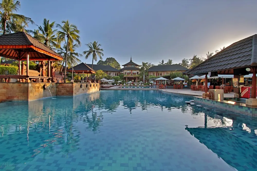 A large swimming pool with a tiled floor extends toward a thatched-roof pavilion on the left, with palm trees behind and an open-air bar and dining area on the right.