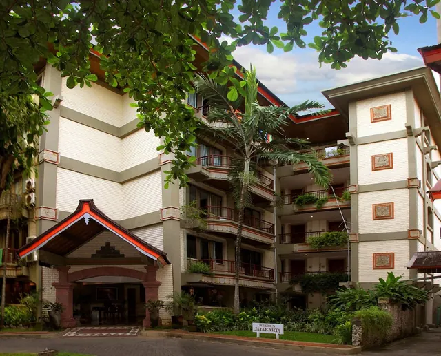 The exterior of a cream-walled, multi-storey hotel building with orange-trimmed roof edges, balconies, a palm tree and a covered entrance, with a sign reading Resident Jayakarta at the front.