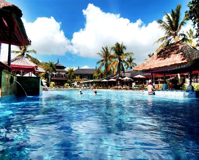A large swimming pool with three people in the water, set among thatched pavilions, palm trees, and sun loungers under umbrellas.
