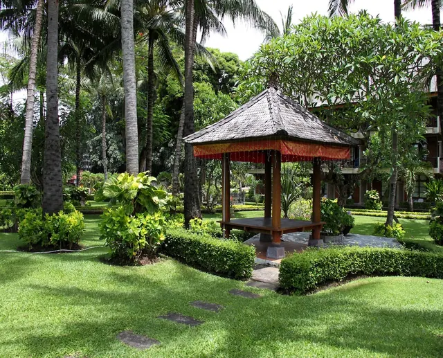 A traditional Balinese wooden pavilion with a shingled roof stands on a trimmed lawn among palm trees, frangipani and hedges, with a hotel building behind it.