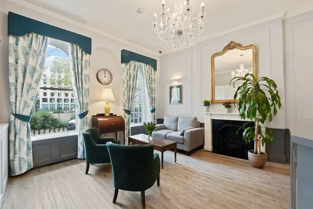 A furnished sitting room with a chandelier, two green armchairs, a grey sofa, a fireplace and tall windows looking onto a street.