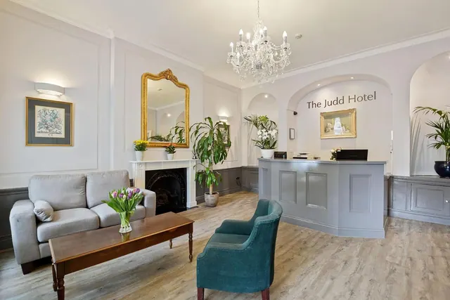 A hotel lobby with a grey sofa, a wooden coffee table holding tulips, a teal armchair, and a reception desk beneath a chandelier, with 'The Judd Hotel' lettering on the wall.