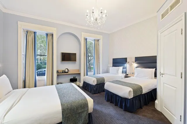 A hotel bedroom with one double bed and two single beds, a chandelier, a wall-mounted television and a kettle on a shelf, with windows showing trees and a street outside.