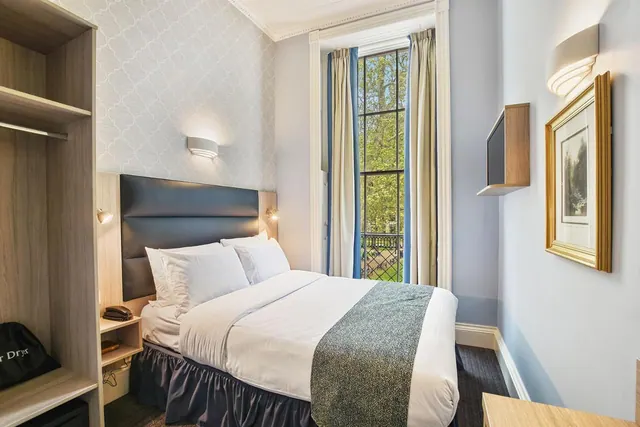 A double bed with white bedding and a dark upholstered headboard sits beside a tall window looking out onto trees, with an open wardrobe on the left.