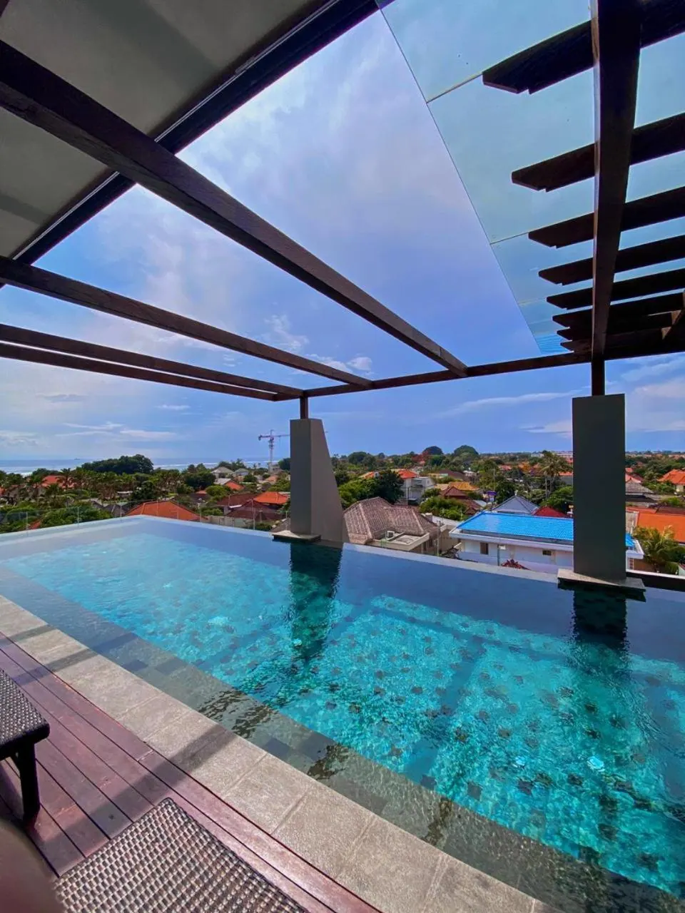 An infinity-edge rooftop pool under a timber pergola, with a view over surrounding houses and trees toward the sea.