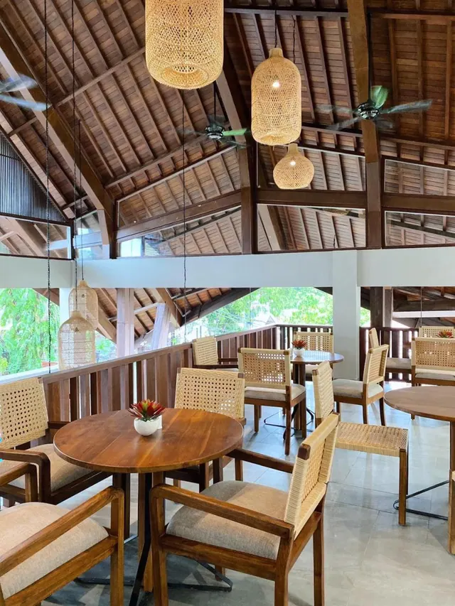 A dining area with wooden tables and woven chairs under rattan pendant lights and a timber-beamed roof, with trees visible beyond the railing.