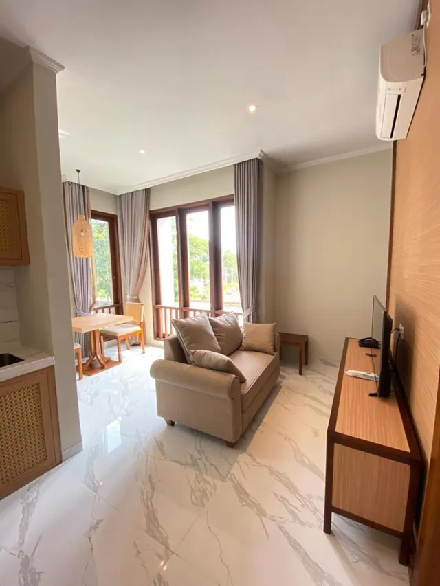 A living area with a beige sofa, a wall-mounted television on a wooden console, a wall air conditioner, a small dining table by the windows, and a marble floor.