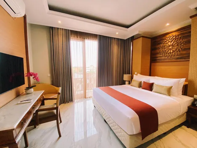 A bedroom with a king bed, a wall-mounted television, a wooden writing desk with a chair, an air conditioning unit, and a glass sliding door opening onto a balcony.