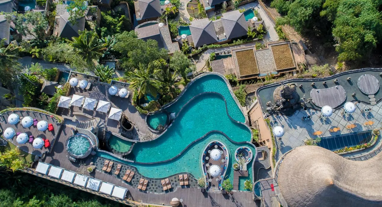 Aerial view of a winding freeform swimming pool surrounded by sun loungers, white umbrellas, a small round jacuzzi and villa roofs among trees.