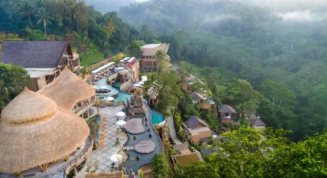 Aerial view of a hillside resort with thatched-roof buildings, a winding pool with white umbrellas, and a misty jungle valley beyond.