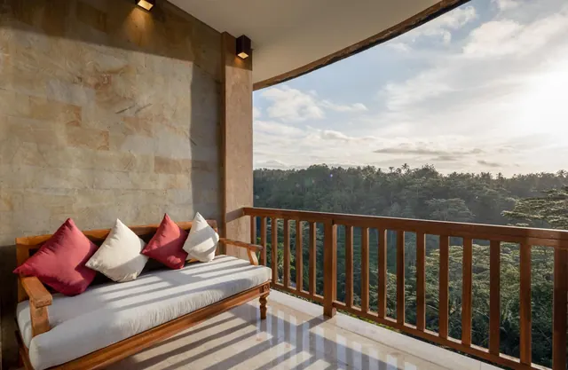 A cushioned daybed with red and white pillows sits on a stone-walled balcony with a wooden railing overlooking a forested valley.