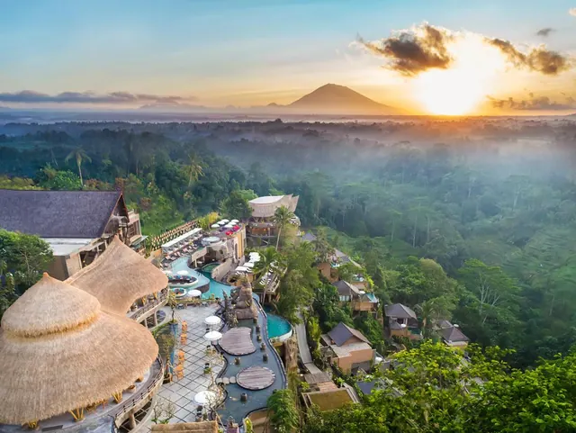 Aerial view of a hotel complex with pools, thatched roofs and jungle below a volcano at sunrise.