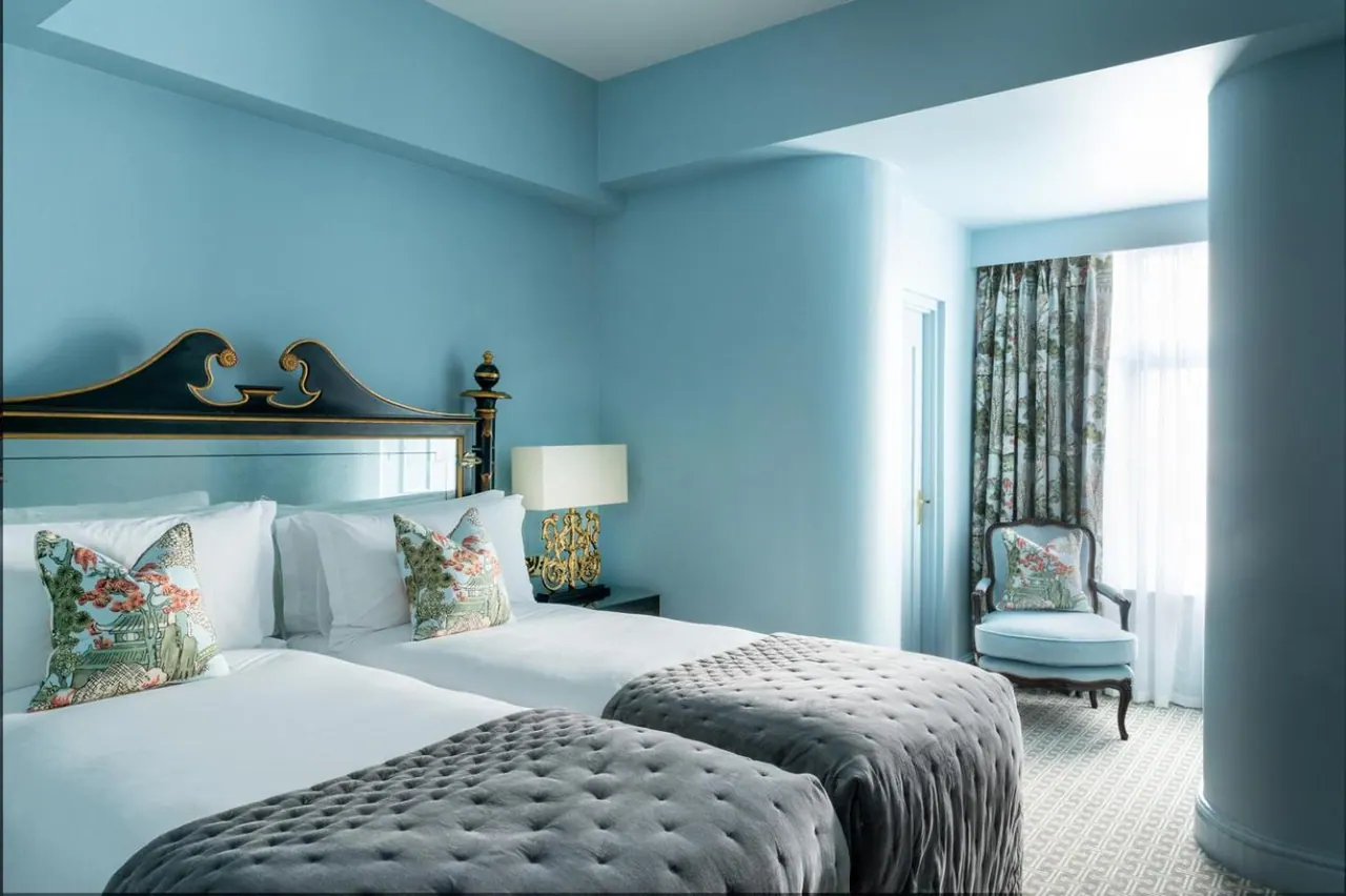 A bedroom with two single beds with grey quilted covers, blue walls, a gold table lamp, and a blue armchair beside a window with sheer curtains.