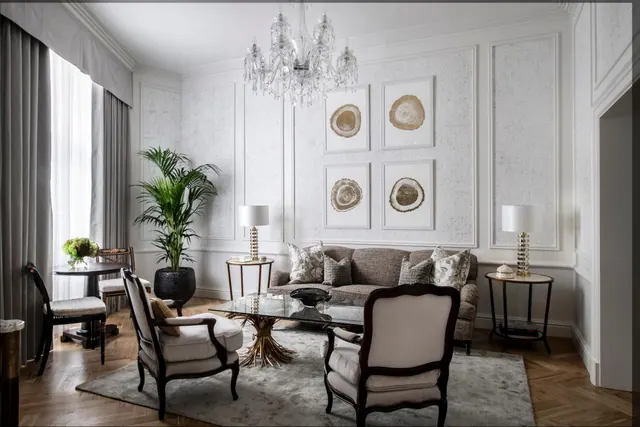 A formal living room with a sofa, two armchairs, a glass coffee table, a crystal chandelier and framed art on a panelled wall.