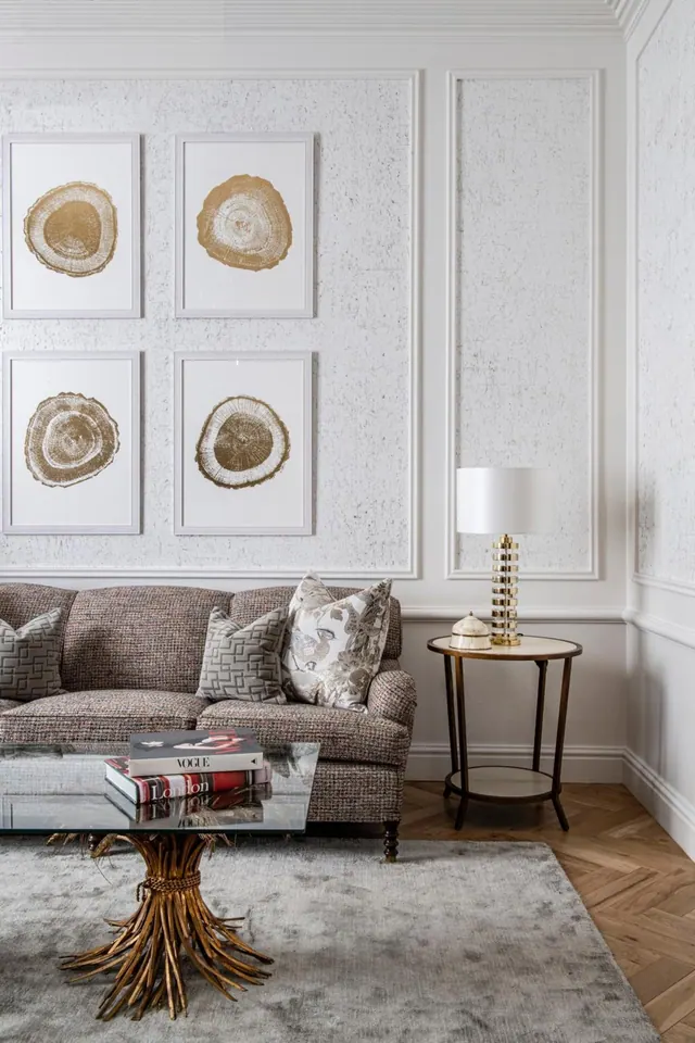 A living area with an upholstered sofa and patterned cushions, a glass coffee table with books, a side table with a lamp, and four framed gold prints on the wall.