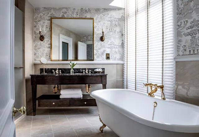 A freestanding clawfoot bathtub with gold fittings sits beside a dark wood vanity with a marble top, below white window blinds and patterned wallpaper.