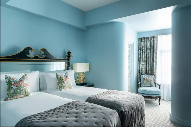 A bedroom with two beds with grey quilted throws, blue walls, a gold table lamp, and an armchair beside a window with sheer curtains.