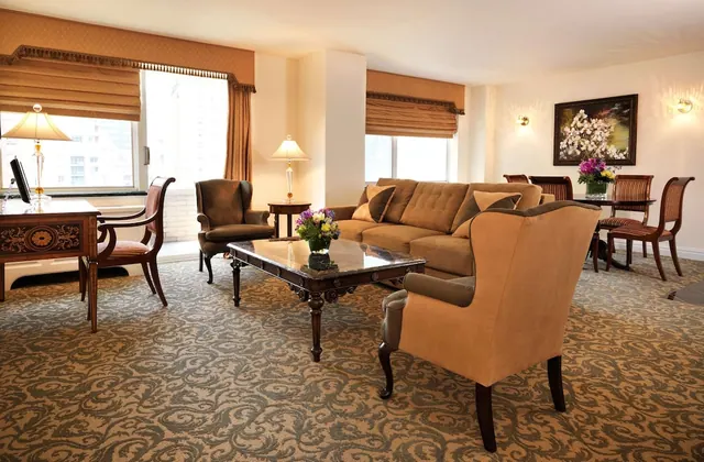 A furnished suite living area with a tufted sofa, wingback armchairs, a marble-topped coffee table, a desk by the window, and a dining table in the background.
