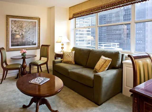 A hotel living area with an olive sofa, a round wooden coffee table, a framed floral print and a large window showing city high-rise buildings.