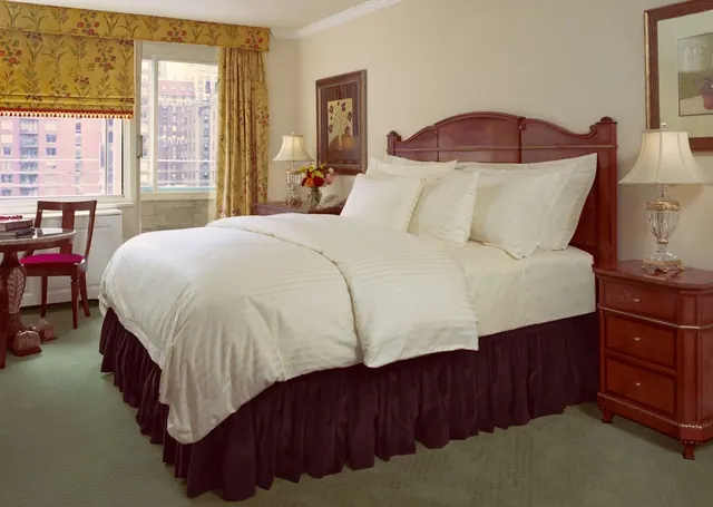 A made king bed with cream striped bedding sits between two wooden nightstands with table lamps, beside a window with yellow floral curtains that looks out onto brick buildings.