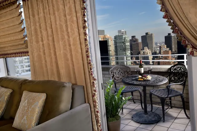 A furnished living room with a gold sofa and tasselled curtains opens onto a balcony with a metal table, two chairs and a city skyline view.