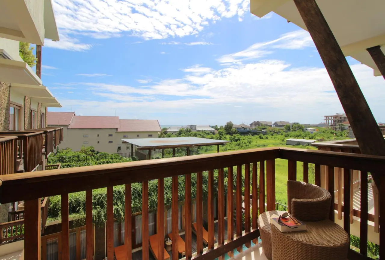 A wooden balcony with two wicker chairs, a small round table holding a book, and a view over green fields and low buildings under a partly cloudy sky.
