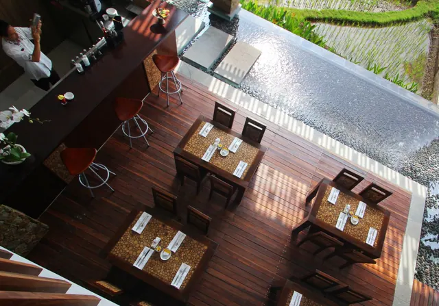 Overhead view of an open-air restaurant with set wooden tables, a bar with stools, a staff member at the counter, and a water feature with rice fields beyond.