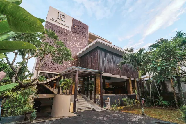 The front of a hotel building with a carved stone panel, a slatted wooden facade, a covered entrance with steps, and tropical plants in the foreground.