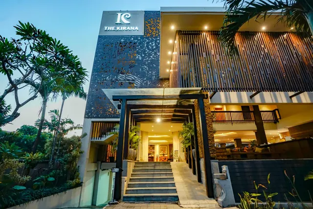 The front of The Kirana hotel at dusk, showing its name sign, a lit entrance with steps leading up to the lobby, and frangipani trees on the left.
