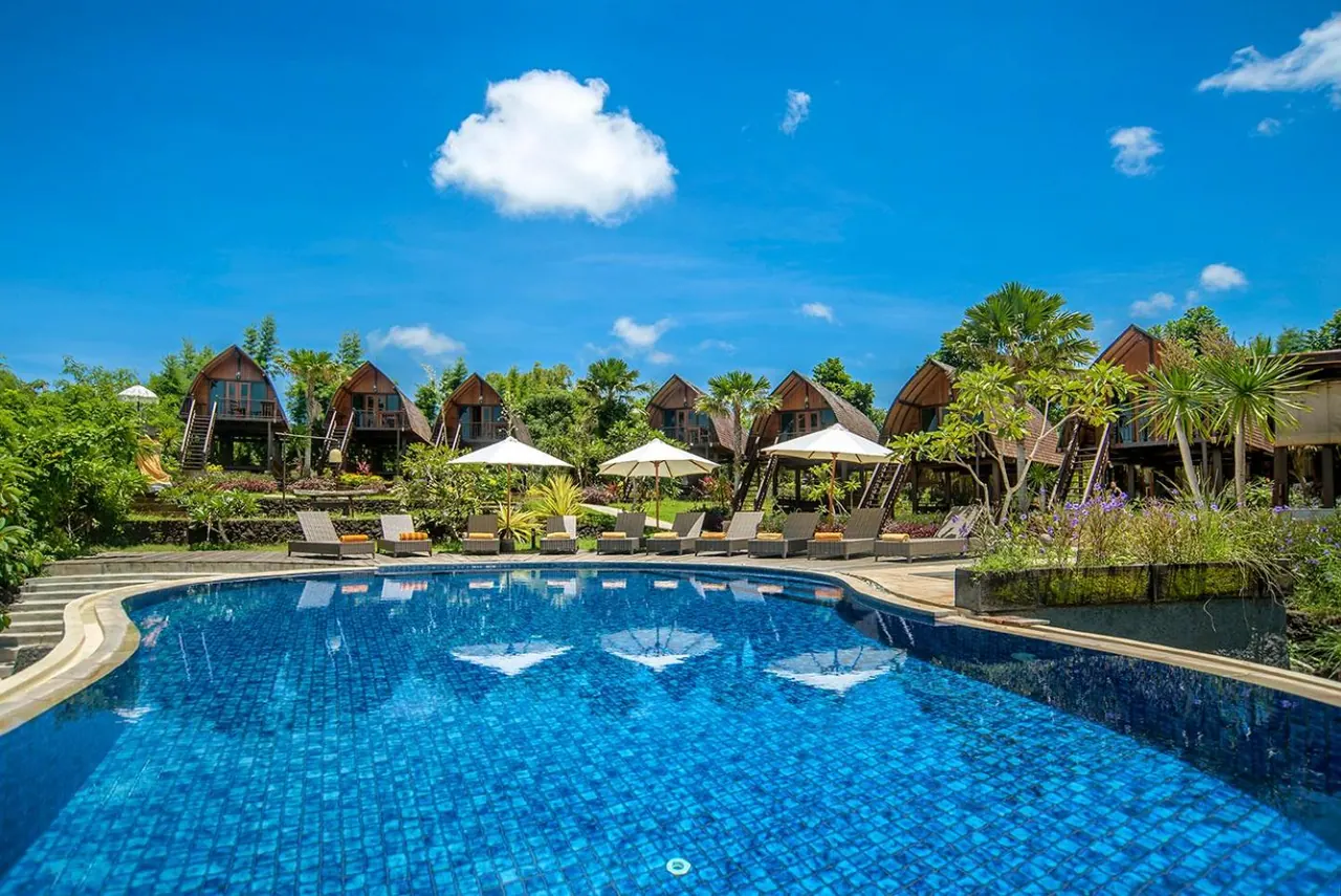 A large blue-tiled swimming pool with sun loungers and umbrellas in front of wooden bungalows and green trees under a blue sky.