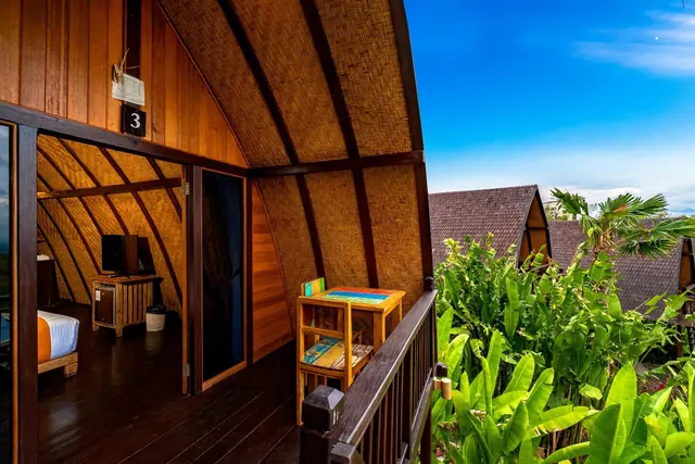 A wooden balcony with a painted table and chair outside a thatched-roof bungalow, with tropical plants and other bungalows beyond the railing.