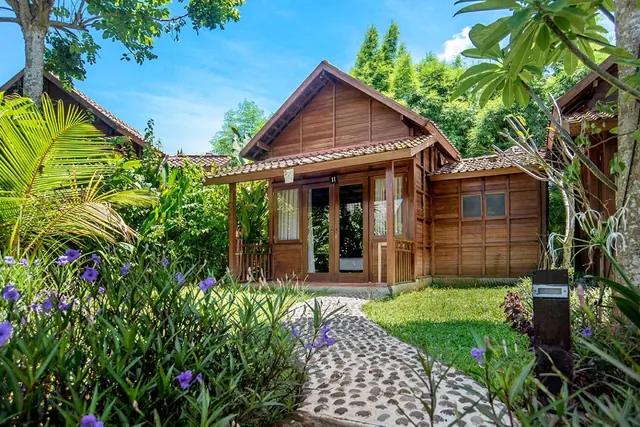 A wooden bungalow with a tiled roof and glass doors stands beside a cobblestone path, lawn and purple flowering plants.