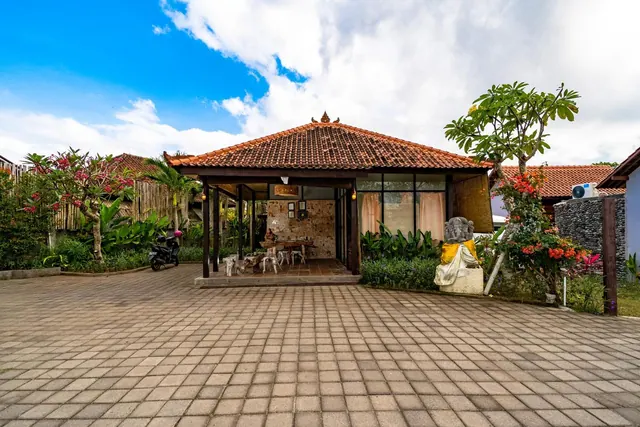 The photo shows the front of a single-storey Balinese-style building with a terracotta tiled roof, a paved forecourt, a parked scooter, frangipani trees and a stone statue beside the entrance.